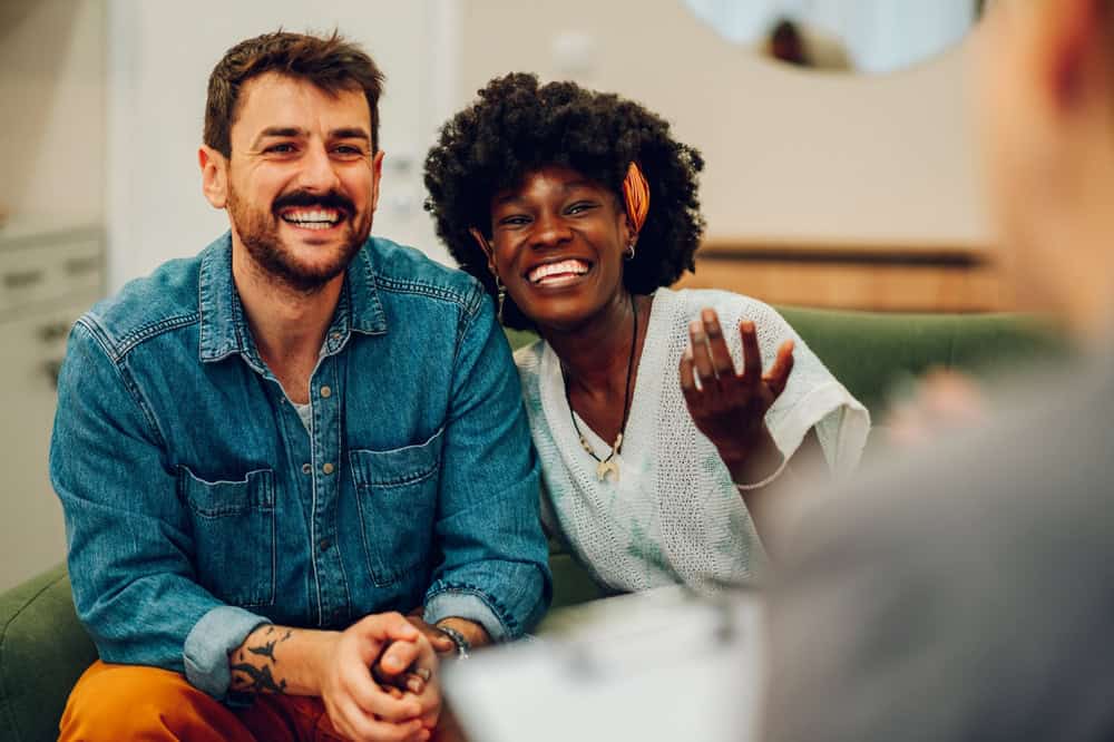 A smiling couple sits on a green couch, engaging in a conversation with an unseen person. The man, with short brown hair and a beard, wears a denim shirt. The woman, with a natural afro and an orange hair accessory, wears a white top. They are clearly benefiting from family counseling in Fort Bend County.