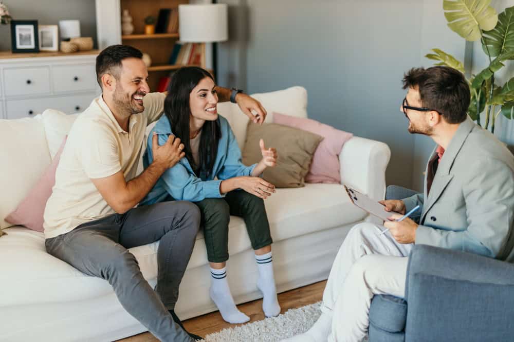 A smiling couple sits on a couch, speaking with a man holding a clipboard who is sitting across from them in an armchair. The room is cozy, with bookshelves, plants, and a dresser in the background, suggesting a relaxed, informal meeting or counseling session.